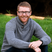 A smiling man with glasses, wearing a grey sweater, sitting outdoors on the grass.
