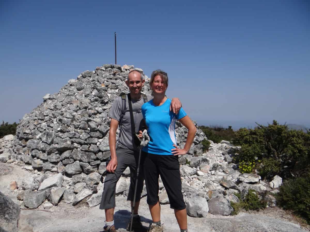 Two hikers smiling at the summit with a large cairn of rocks under a clear blue sky.
