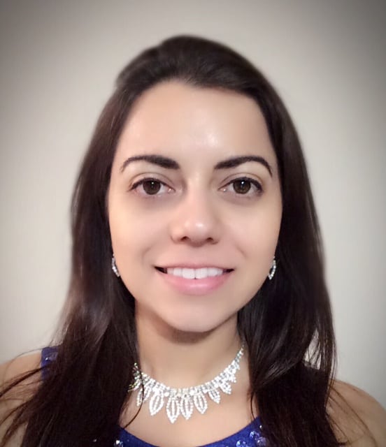 A woman with long dark hair and a friendly smile wearing sparkling earrings and a necklace with a blue attire, against a neutral background.