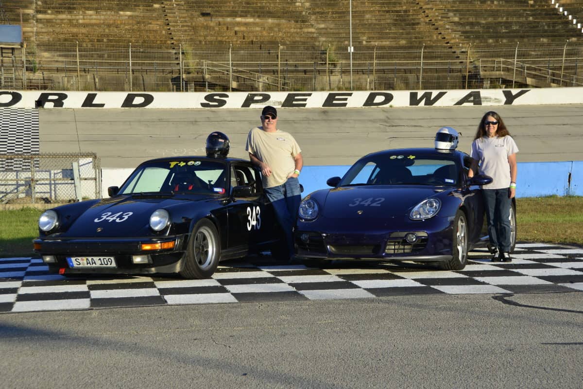 Two people standing by their racing cars on a speedway track, exuding a sense of pride and readiness for competition.