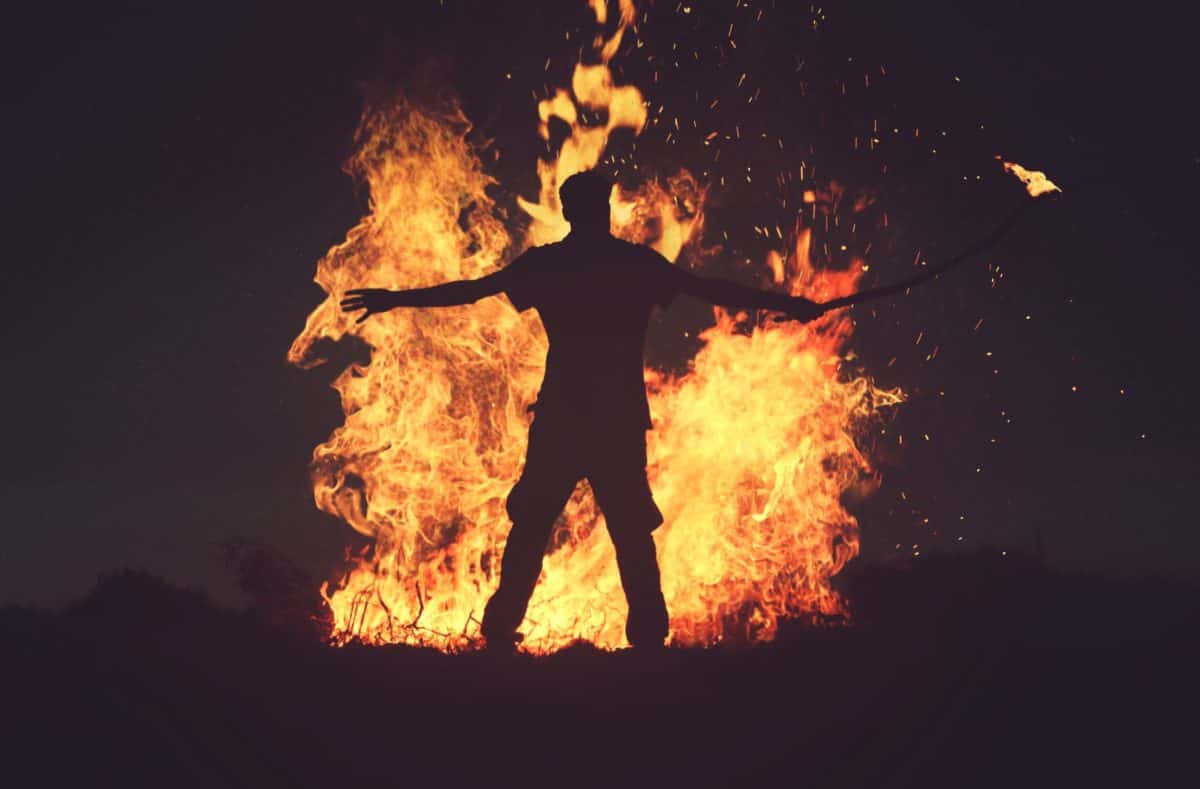 A silhouetted figure stands before a large, intense bonfire, spreading their arms amidst the fiery blaze in a dramatic and powerful stance.