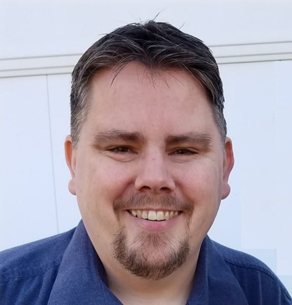 A smiling person with short hair and a goatee standing in front of a plain background.