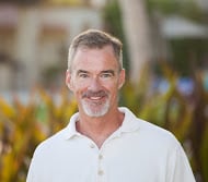 Middle-aged man with a friendly smile, wearing a white shirt, standing in front of a blurred background with greenery.