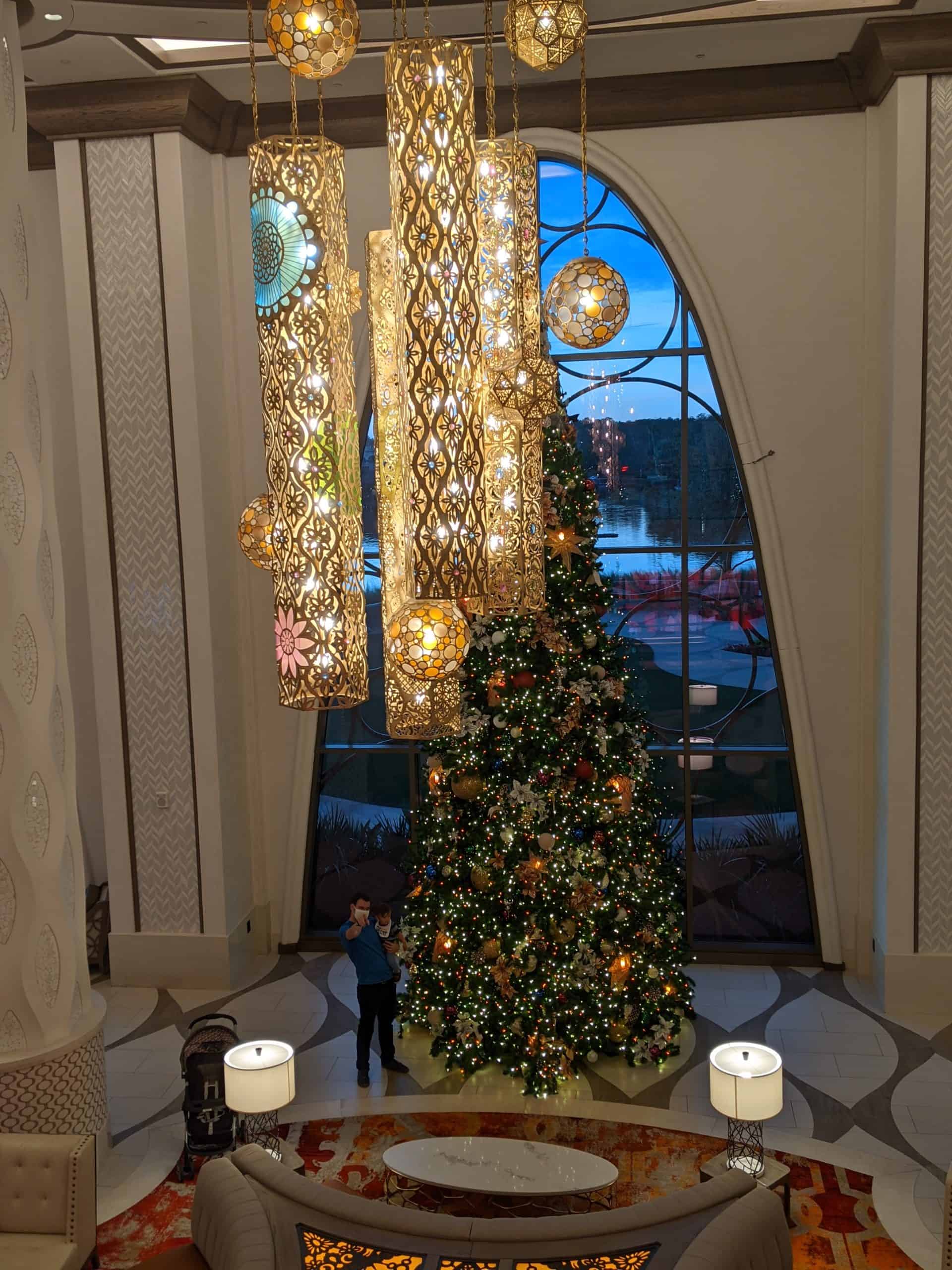 A child stands in awe before a towering christmas tree, adorned with twinkling lights and festive decorations, in an elegant lobby with ornate hanging lanterns casting a warm, inviting glow.