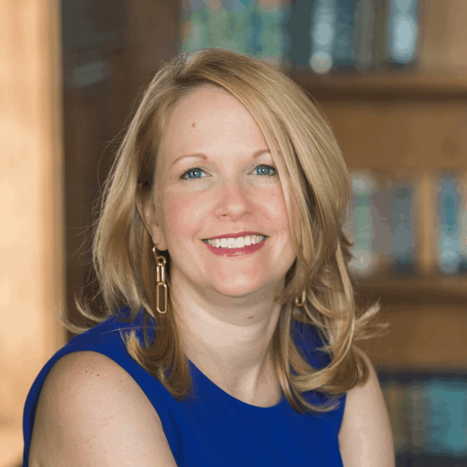 A smiling woman with shoulder-length blonde hair, wearing a blue top and gold earrings, posing for a professional portrait with a soft-focus background.