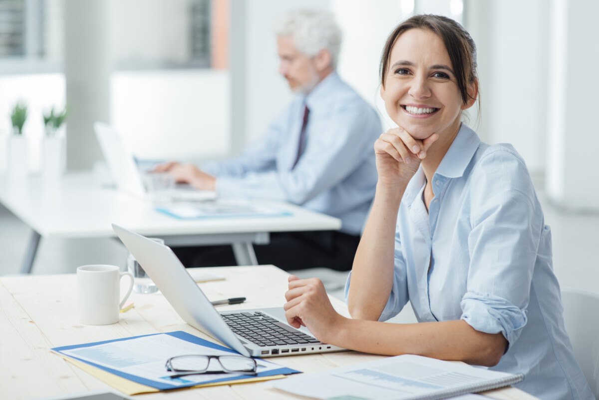 Smiling young technology worker sitting at office desk and working with a laptop, she is looking at camera