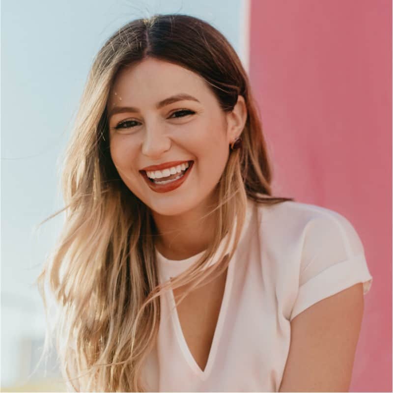 A cheerful young woman smiling brightly, with her hair gently tousled by the breeze, against a softly blurred pink background.
