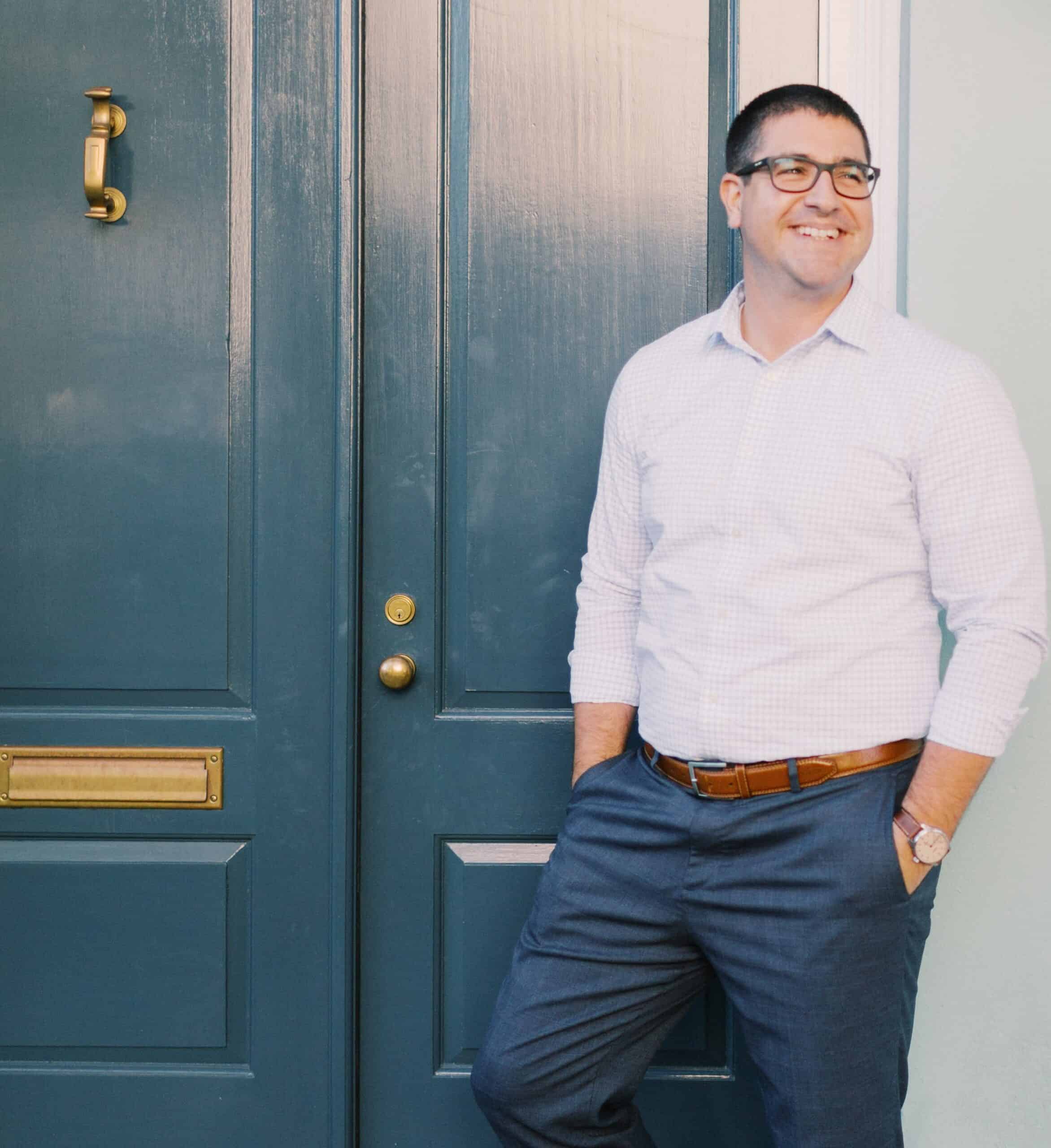 A man in a button-up shirt and glasses standing with a confident smile beside a dark blue door with brass fixtures.