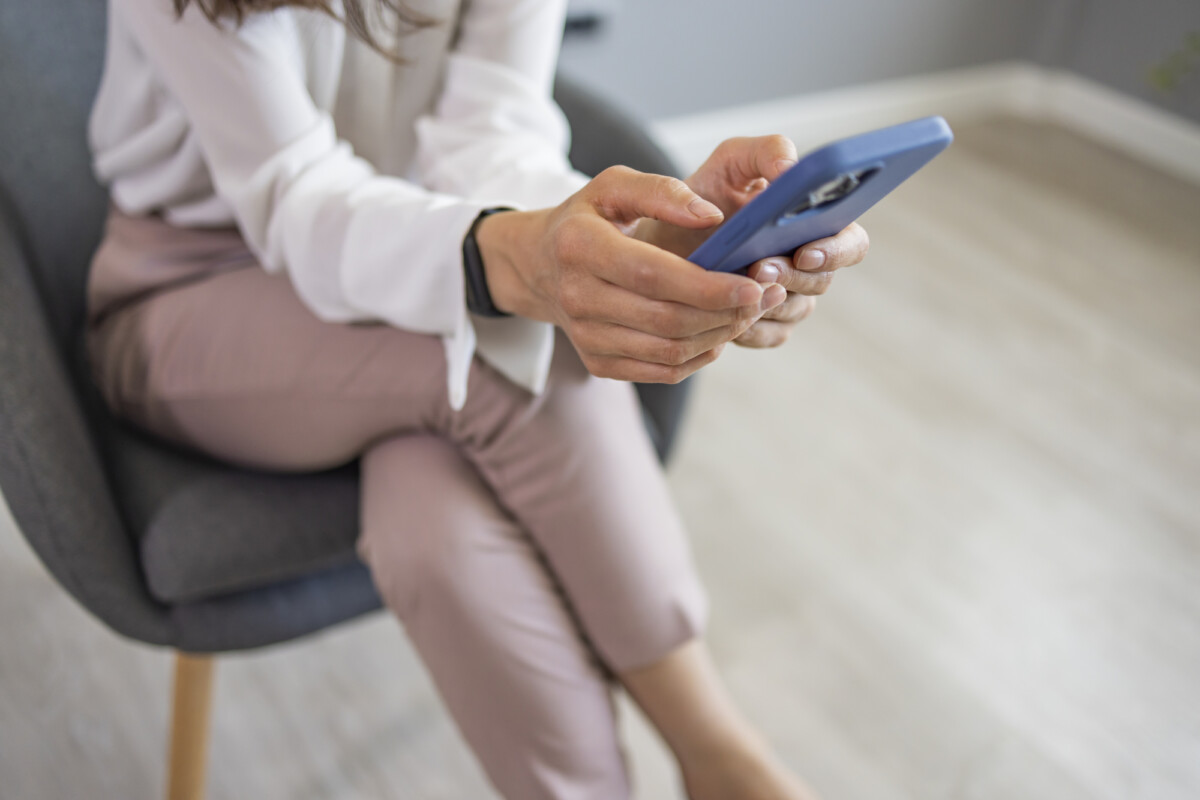 A person seated in a modern chair, casually dressed in a white shirt and pink pants, scrolling through a smartphone with focused attention.