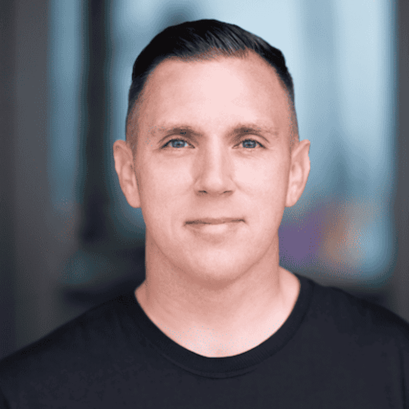 Confident man with a neat haircut smiling slightly at the camera, wearing a black crew neck t-shirt against a blurred background.