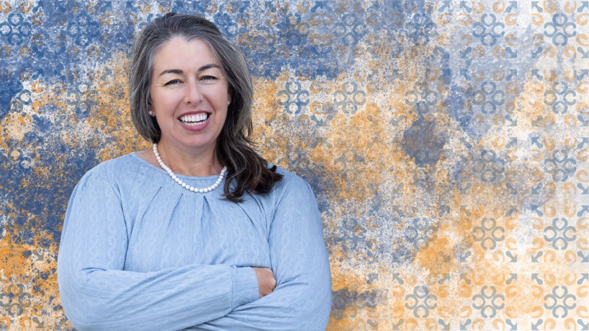 A smiling woman with long gray hair, wearing a light blue blouse and a pearl necklace, stands with her arms crossed against a blue and orange textured patterned background.