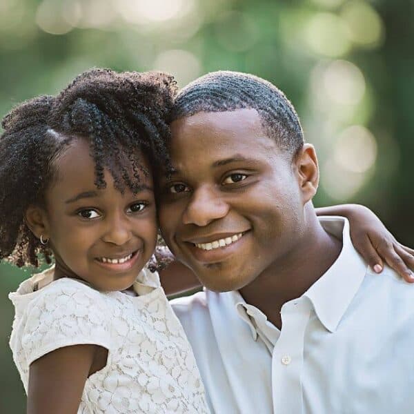 A smiling man and young girl pose together against a blurred green background. The girl, with curly hair, wears a white dress and has her arm around the man's shoulder. The man is wearing a white shirt. Both appear happy and content.