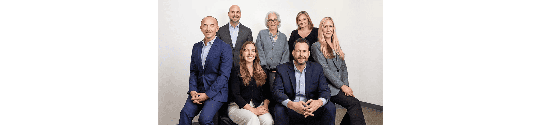 Seven people in business attire are posed together in a group. Four are seated in front, and three are standing behind them against a plain white background. They are all smiling at the camera.