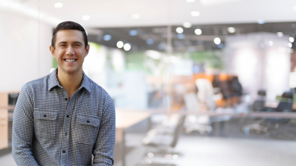 A man in a checkered shirt smiles while sitting in a modern office with glass walls, blurred chairs, and desks in the background.