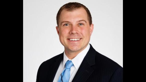 A man in a dark suit, light blue tie, and white shirt smiles at the camera against a plain white background.