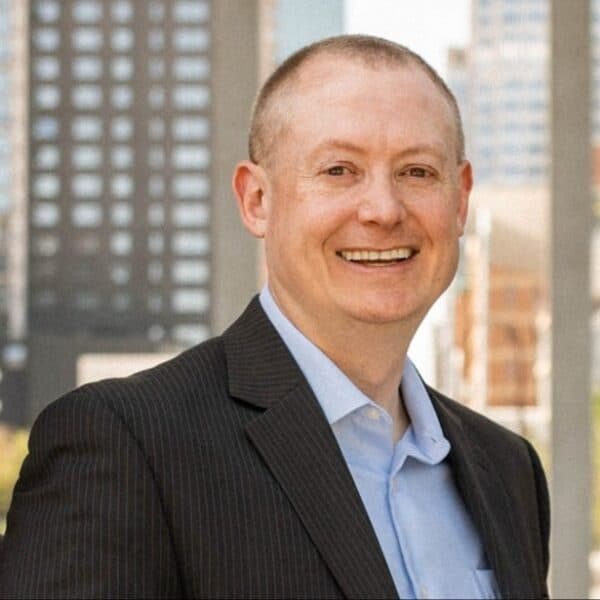 A smiling man in a dark striped suit jacket and light blue dress shirt stands outdoors in front of tall, blurred city buildings.