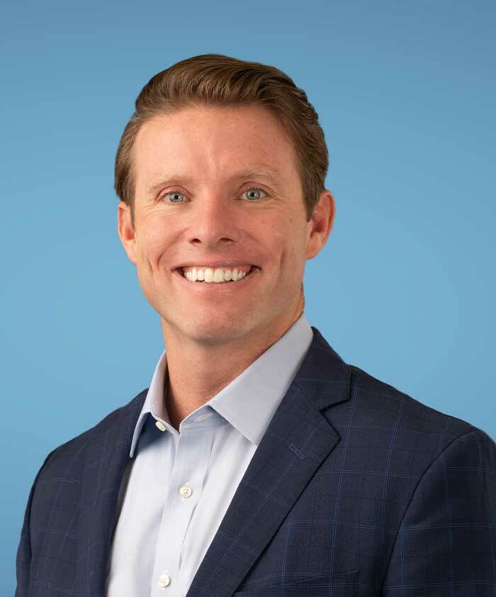 A man with light brown hair wearing a blue suit jacket and a light blue dress shirt smiles in front of a plain blue background.