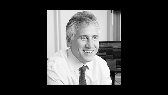 A middle-aged man with gray hair, wearing a dress shirt and tie, smiles while sitting at a desk. A computer monitor is visible in the background. The image is black and white.