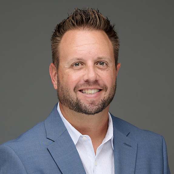 A man with short, spiked brown hair and a trimmed beard smiles at the camera. He is wearing a light blue blazer over a white collared shirt, with a neutral gray background.