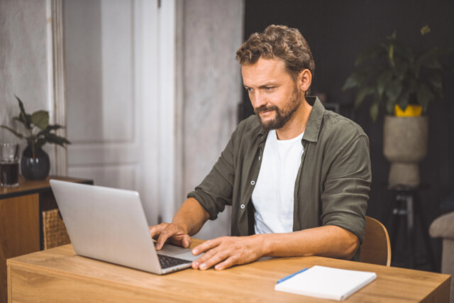 A man with short brown hair and a beard sits at a wooden desk using a laptop, with a notebook nearby. He appears focused, and there are plants and a glass of water in the background.