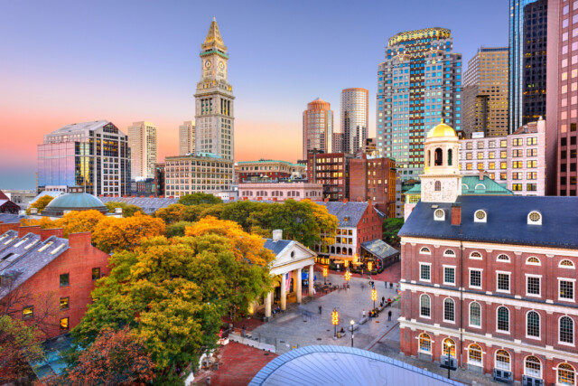 Boston cityscape at sunset with historic Faneuil Hall and Quincy Market in the foreground, trees with autumn foliage, and modern skyscrapers in the background under a colorful sky.