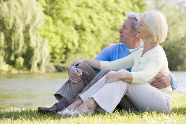 An older couple sits on grass by a lake, holding each other and looking into the distance, surrounded by lush green trees on a sunny day.