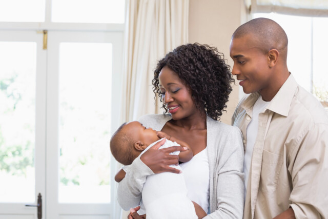 A smiling couple stands indoors, lovingly holding and looking at their baby. The mother cradles the baby in her arms while the father stands close, both appearing happy and engaged with the child.