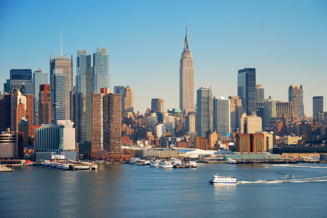 A view of the New York City skyline with the Empire State Building and other skyscrapers, seen across the Hudson River with several boats and ferries on the water under a clear blue sky.