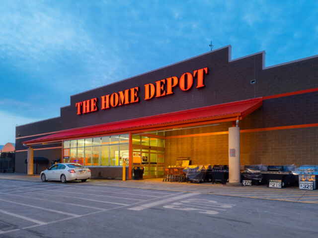 The Home Depot store exterior at sunset, with bright orange sign, parked white car, shopping carts, and outdoor merchandise near the entrance.