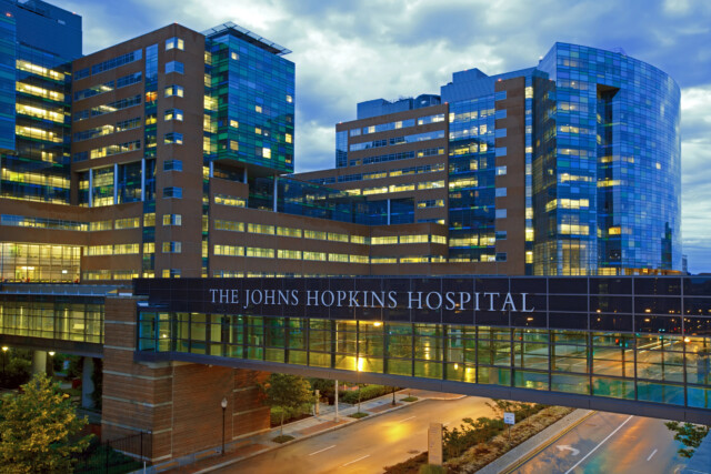 Modern glass and brick hospital building at dusk, with illuminated windows and a skybridge labeled "The Johns Hopkins Hospital" over a quiet street.