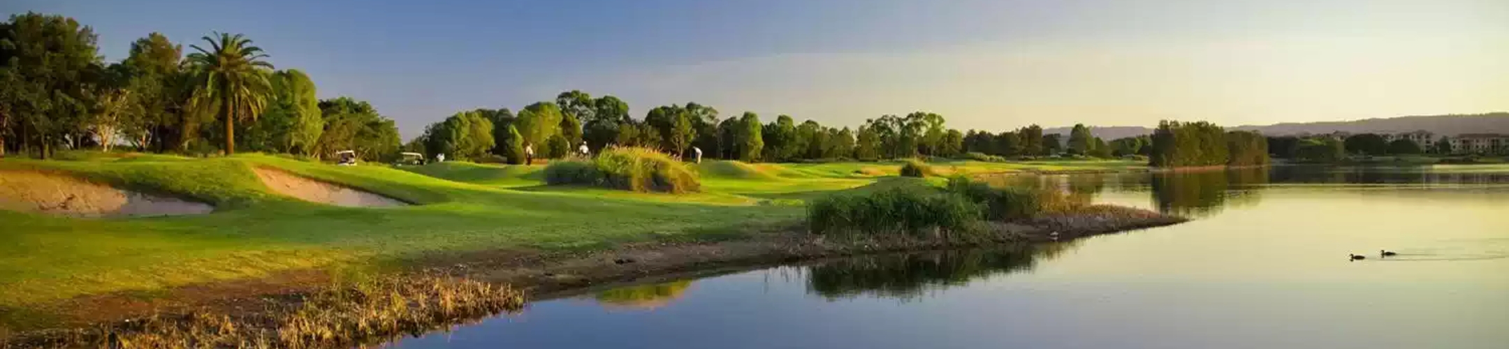 A scenic golf course with lush green fairways, sand bunkers, and scattered trees beside a calm lake, with a clear sky and distant hills in the background.