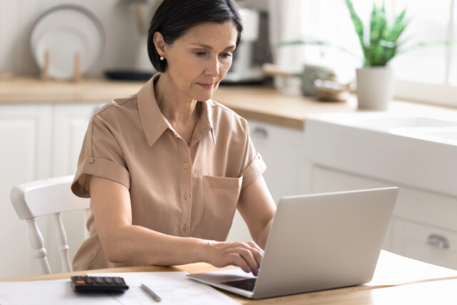 A woman with short dark hair is sitting at a kitchen table, working on a laptop. Papers, a pen, and a calculator are on the table. She appears focused, and there is a plant and kitchenware in the background.