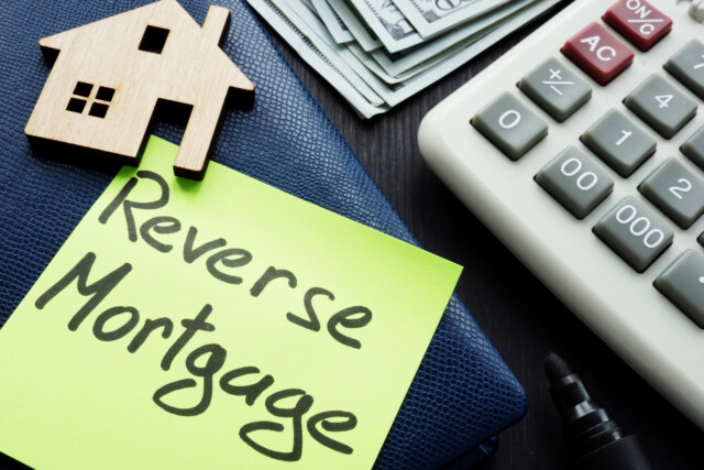 A sticky note labeled "Reverse Mortgage" sits on a blue wallet next to a wooden house figure, a stack of dollar bills, a calculator, and a black marker.