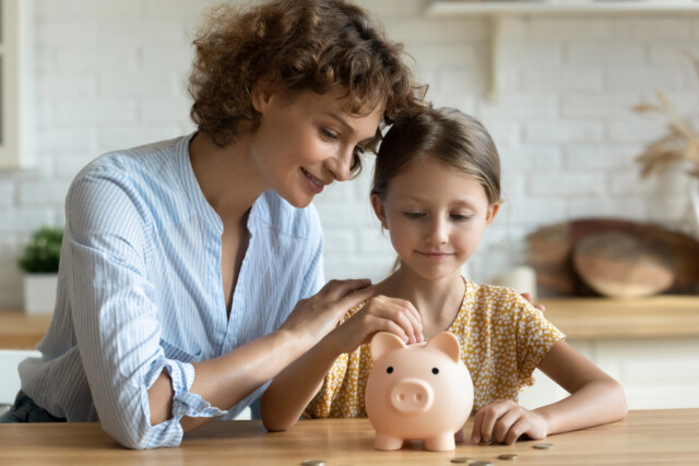 A woman and a young girl smile while putting coins into a pink piggy bank on a table, teaching about saving money in a bright kitchen.